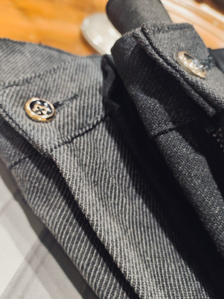 Nahaufnahme einer dunkelgrauen Hose mit feiner Struktur; Fokus auf Knopf, Gürtelriemendetail und dem hochwertigen, leicht elastischen Stoff.