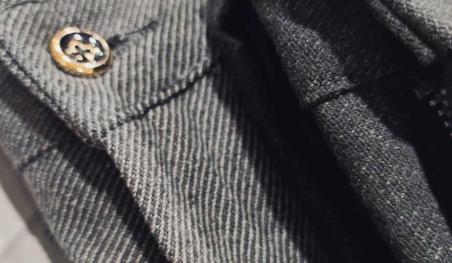 Nahaufnahme einer dunkelgrauen Hose mit feiner Struktur; Fokus auf Knopf, Gürtelriemendetail und dem hochwertigen, leicht elastischen Stoff.