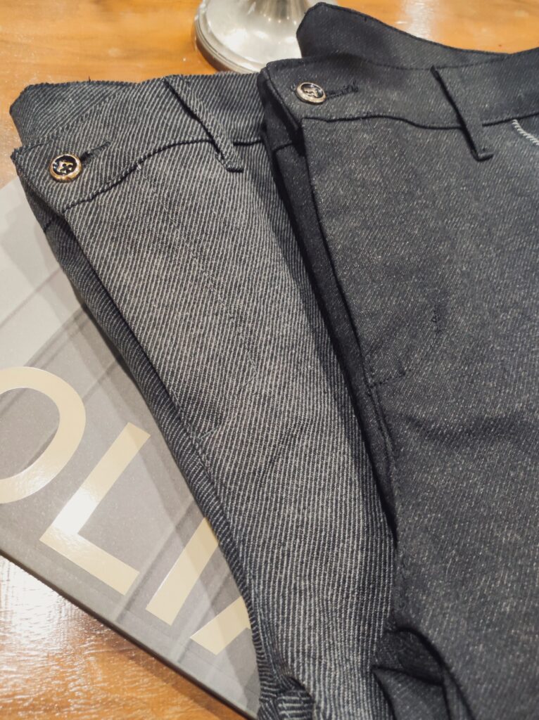 Nahaufnahme einer dunkelgrauen Hose mit feiner Struktur; Fokus auf Knopf, Gürtelriemendetail und dem hochwertigen, leicht elastischen Stoff.