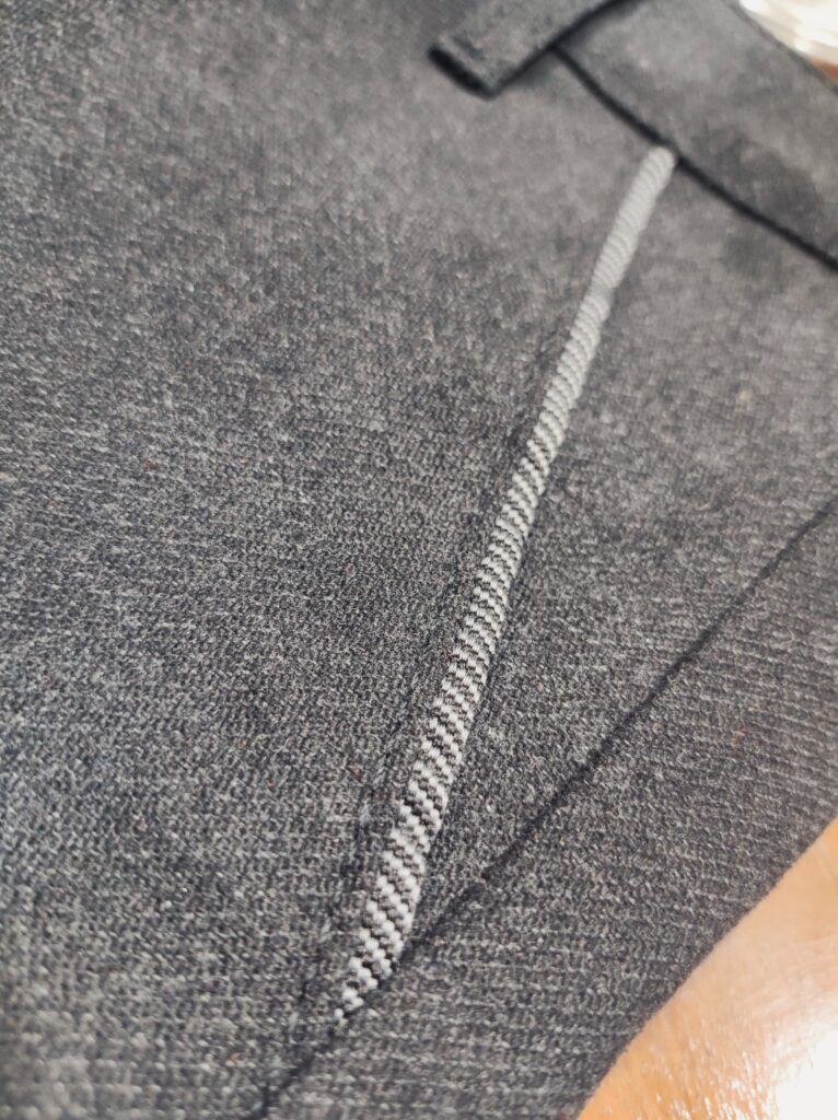 Nahaufnahme einer dunkelgrauen Hose mit feiner Struktur; Fokus auf Knopf, Gürtelriemendetail und dem hochwertigen, leicht elastischen Stoff.